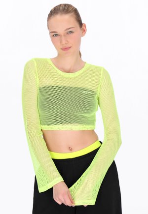 Junge Frau trägt ein neon-gelbes, langärmeliges, durchsichtiges Crop-Top über einem schwarzen Sport-BH und schwarze Hosen mit neon-gelbem Taillenbund.