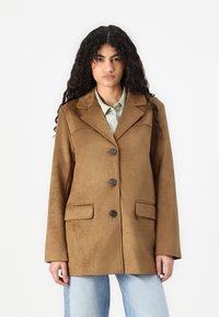 Brown suede blazer with a notched collar, three buttons, and two front pockets. Soft texture, relaxed fit, paired with a light shirt.