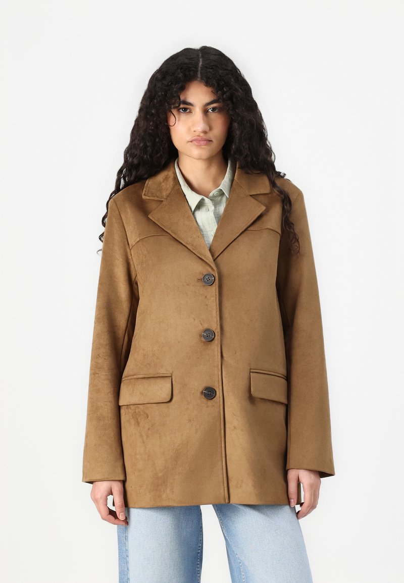 Brown suede blazer with a notched collar, three buttons, and two front pockets. Soft texture, relaxed fit, paired with a light shirt.