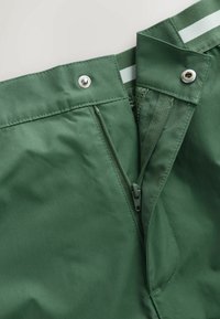 Ceinture de pantalon verte avec une fermeture éclair partiellement ouverte et deux boutons-pression argentés, avec une bande élastique intérieure verte et blanche à rayures.