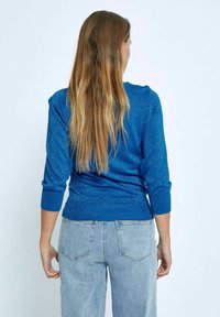 Modra, oprijeta pulover s tremi četrtinami rokavov, teksturirana z bleščicami. V kombinaciji s svetlim denimom, prikazana z zadnje strani.