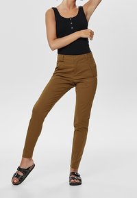 Brown slim-fit trousers with front pockets, paired with a black sleeveless top and black sandals featuring buckled accents.