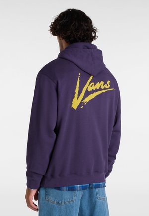 Mężczyzna ubrany w ciemnofioletową bluzę z kapturem z dużym żółtym logo "Vans" na plecach, w połączeniu z niebieskimi dżinsami i niebieską koszulą w kratę pod spodem.