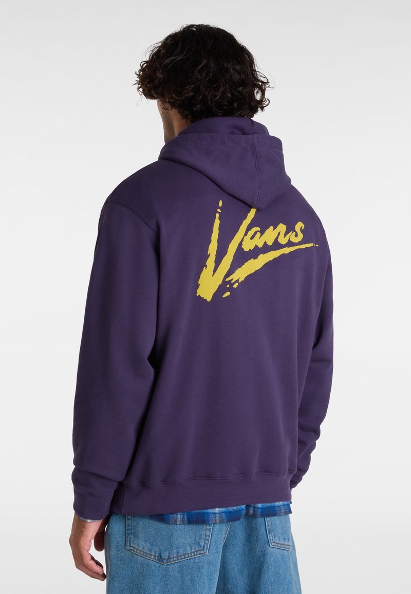 Homme portant un sweat à capuche violet foncé avec un grand logo jaune "Vans" au dos, associé à un jean bleu et une chemise à carreaux bleue en dessous.