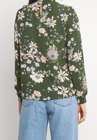 Blommig blus i mörkgrön med långa ärmar, utrustad med plisserade manschetter och en liten slits bak, dekorerad med rosa, vita och gula blommor.