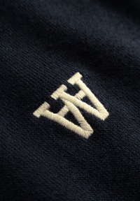 Marinefarbener Stoff mit einem auffälligen, cremefarbenen gestickten Logo, das ein geschichtetes geometrisches Design mit einer weichen, strukturierten Oberfläche zeigt.