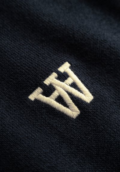 Textil marino con un destacado logotipo bordado color crema, que presenta un diseño geométrico en capas con un acabado suave y texturizado.