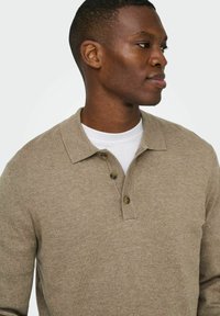 Polo de punto beige con cierre de tres botones, tela texturizada y cuello clásico, sobre una camiseta blanca de cuello redondo.