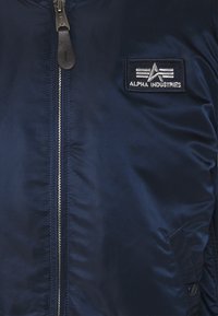 Jachetă de bombardier de culoare bleumarin, cu o finisare netedă, fermoar central și un patch brodat Alpha Industries pe partea stângă a pieptului.