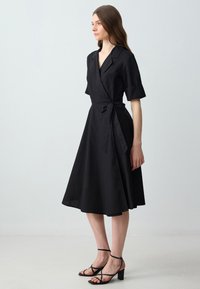 Femme portant une robe noire à manches courtes et à nouer à la taille, associée à des sandales à talons blocs noires à lanières, se tenant de profil.
