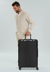 Hombre con camisa beige sosteniendo el asa de una gran maleta rígida negra con ranuras diagonales sobre un fondo blanco.