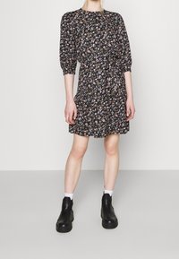 Vrouw in een zwarte bloemenjurk met roze en witte bloemen, witte sokken en zwarte enkellaarzen, staande tegen een effen achtergrond.