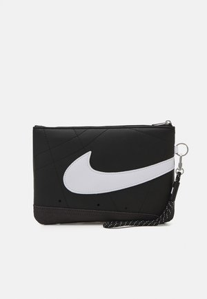 Pochette noire zippée avec grand logo swoosh blanc et sangle noire à pois blancs attachée à un porte-clés en métal.