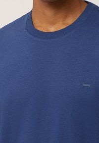 Blå bomuldst-shirt med rund halsudskæring og lille, præget gravhund-logo på brystet, glat tekstur med jævne syninger.