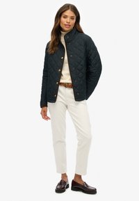 Veste matelassée de couleur vert foncé avec un col montant et des boutons dorés, portée sur un pull crème et un pantalon blanc, accompagnée de mocassins noirs.