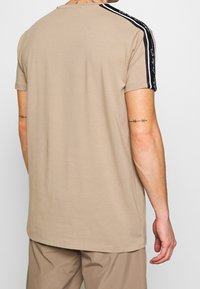 Béžové tričko, krátké rukávy, kulatý výstřih, s černými bočními pruhy a bílým textem. Měkká tkanina, volný střih, rovný lem.