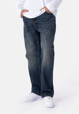 Person iført løse blå jeans, hvid langærmet skjorte og hvide sneakers, står med hænderne i lommerne mod en ensfarvet baggrund.