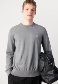 Grauer Strickpullover mit rundem Halsausschnitt, gerippten Bündchen und Saum, mit einem kleinen Logo-Patch auf der Brust; kombiniert mit einem schwarzen Leder-Rucksack.