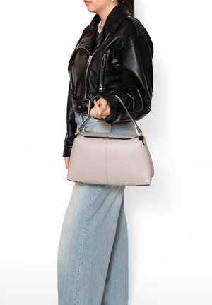 Donna con giacca di pelle nera e jeans azzurri chiari che tiene una borsa beige per il manico al suo fianco.