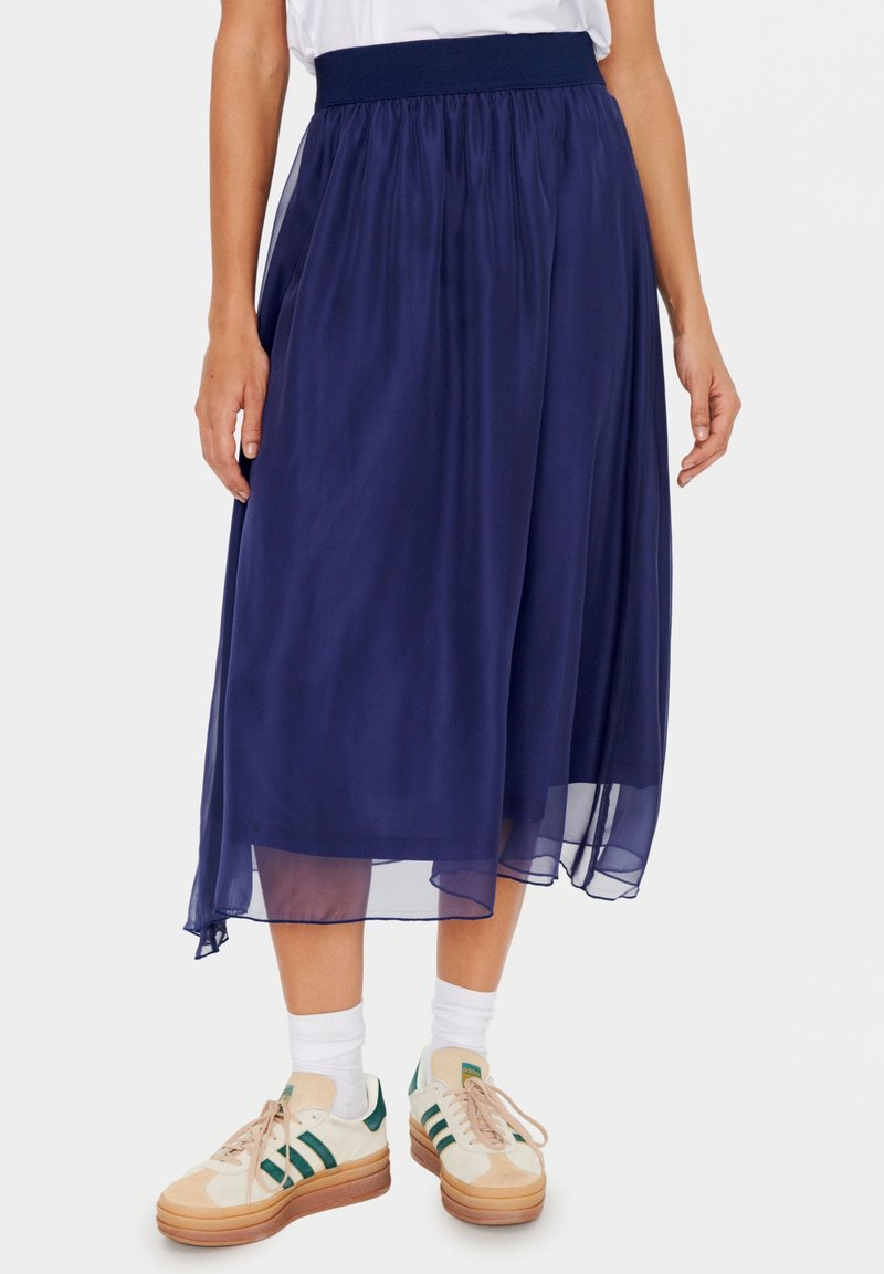 Sininen midi-hame, joka on valmistettu läpikuultavasta kankaasta, jossa on joustava vyötärö ja virtaava siluetti, yhdistettynä vaaleanbeigeihin ja vihreisiin lenkkareihin.