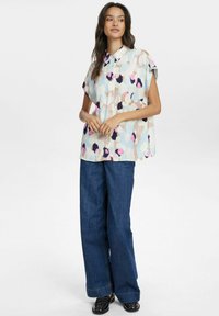 Camicia a maniche corte con bottoni caratterizzata da un motivo astratto colorato con rosa, blu e nero su uno sfondo chiaro, abbinata a pantaloni in denim a gamba larga.