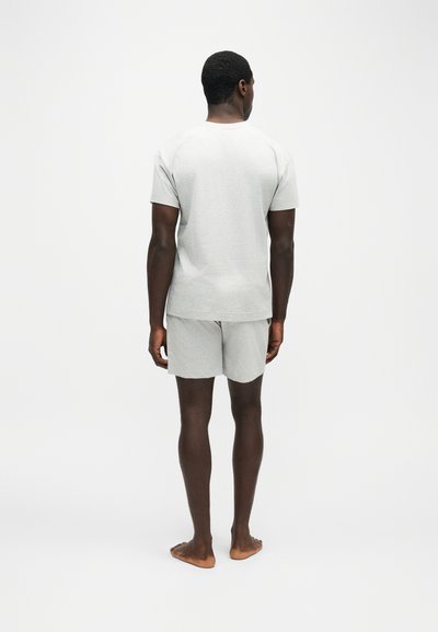 Homme debout pieds nus, de dos, portant une chemise à manches courtes gris clair et un short assorti sur fond blanc uni.