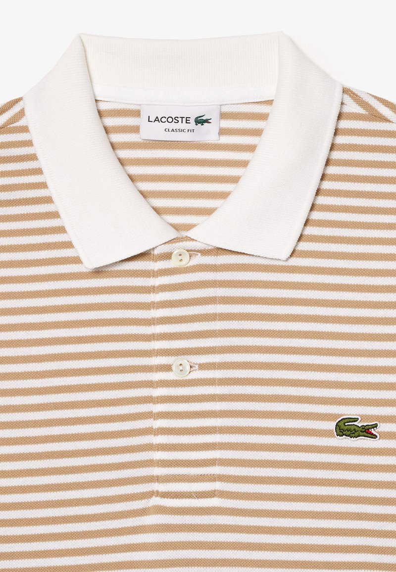 Polo Lacoste à rayures présentant des rayures beiges et blanches, un col blanc et un logo crocodile vert sur la poitrine gauche. Patte de boutonnage à deux boutons.