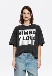 Čierne oversized tričko s kovovým strieborným logom "BIMBA Y LOLA", s krátkymi rukávmi a uvoľneným strihom, skombinované s svetlo modrými džínami.