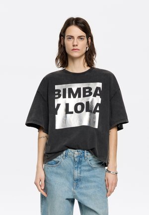 Melnas oversized T-kreklu ar metāliski sudraba "BIMBA Y LOLA" logo, īsām piedurknēm un brīvu piegriezumu, pārī ar gaiši ziliem džinsiem.