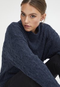 Pull oversized en maille texturée bleu marine avec un col haut et décontracté et des manches larges, porté avec un pantalon ajusté noir.