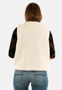Gilet in pile di colore crema, morbido e senza maniche, con colletto alto, abbinato a una blusa nera trasparente a fiori e jeans blu.