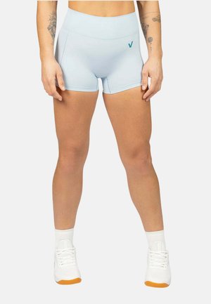 Persona con pantalones cortos deportivos ajustados de color azul claro, calcetines blancos y zapatillas deportivas blancas, de pie frente a un fondo liso.
