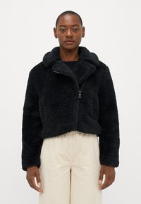 Casaco cropped de pelúcia sintética preta com um grande colarinho, fecho de zíper frontal e bolsos laterais, combinado com calças de corduroy bege claro.