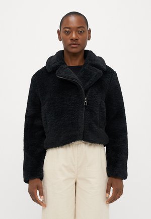 Zwarte gecropte faux fur jas met een grote kraag, ritssluiting aan de voorkant en zijzakken, gecombineerd met lichtbeige corduroy broeken.