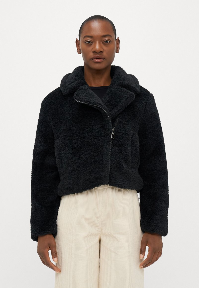 Casaco cropped de pelúcia sintética preta com um grande colarinho, fecho de zíper frontal e bolsos laterais, combinado com calças de corduroy bege claro.