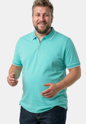 Uśmiechnięty mężczyzna z brodą, ubrany w turkusową koszulkę polo z krótkim rękawem i ciemne dżinsy, stojący na jednolitym jasnym tle.