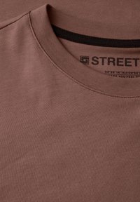 Hnědý bavlněný tričko s kulatým výstřihem, s kontrastním černým vnitřním lemem a potištěnou etiketou. Hladká textura, jednoduchý design.