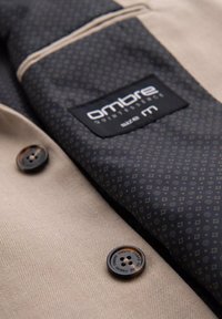 Manteau beige avec deux boutons brun foncé embossés du nom de la marque et une doublure intérieure noire à motifs, taille médium.