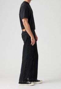 Homme portant un t-shirt noir, un jean noir ample, des baskets noires avec des semelles blanches, debout de profil sur un fond gris uni.