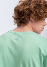 Camicia verde chiaro a maniche corte con un sottile dettaglio grafico a forma di forbici sulla schiena, vicino al colletto, realizzata in un tessuto testurizzato.
