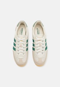 Ténis brancos com riscas verdes, feitos de camurça e couro, com pormenores perfurados e sola de borracha. Marca visível nas palmilhas.