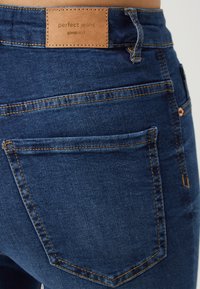 Mörkblå denimjeans med en tan läderlapp märkt "perfekta jeans" nära midjan, med standard bakfickor med synlig sömnad.