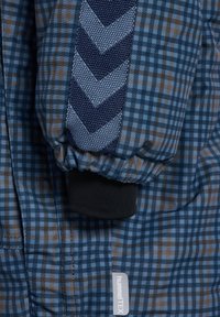 Blaues Karojackett mit einer strukturierten Chevron-Streife in Marineblau, ausgestattet mit elastischen Nähten und einem Logo-Label. Der Stoff hat ein kariertes Muster.