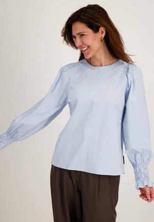 Lachende vrouw met bruin haar draagt een lichtblauwe blouse met lange mouwen en geplooide manchetten, en losse donkerbruine broek, staand tegen een beige achtergrond.