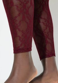 Bordowe koronki legginsy z kwiatowym wzorem, o przejrzystej fakturze i dopasowanym kroju, kończące się na kostce, ukazujące bose stopy poniżej.
