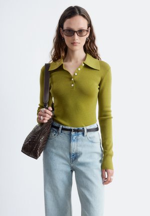 Donna che indossa una camicia verde a maniche lunghe con colletto, jeans azzurri chiari con cintura nera, occhiali da sole scuri e tiene una borsa intrecciata marrone.