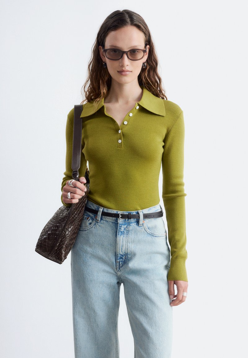 Femme portant une chemise verte à manches longues avec col, un jean bleu clair avec ceinture noire, des lunettes de soleil foncées, et tenant un sac à main tressé marron.