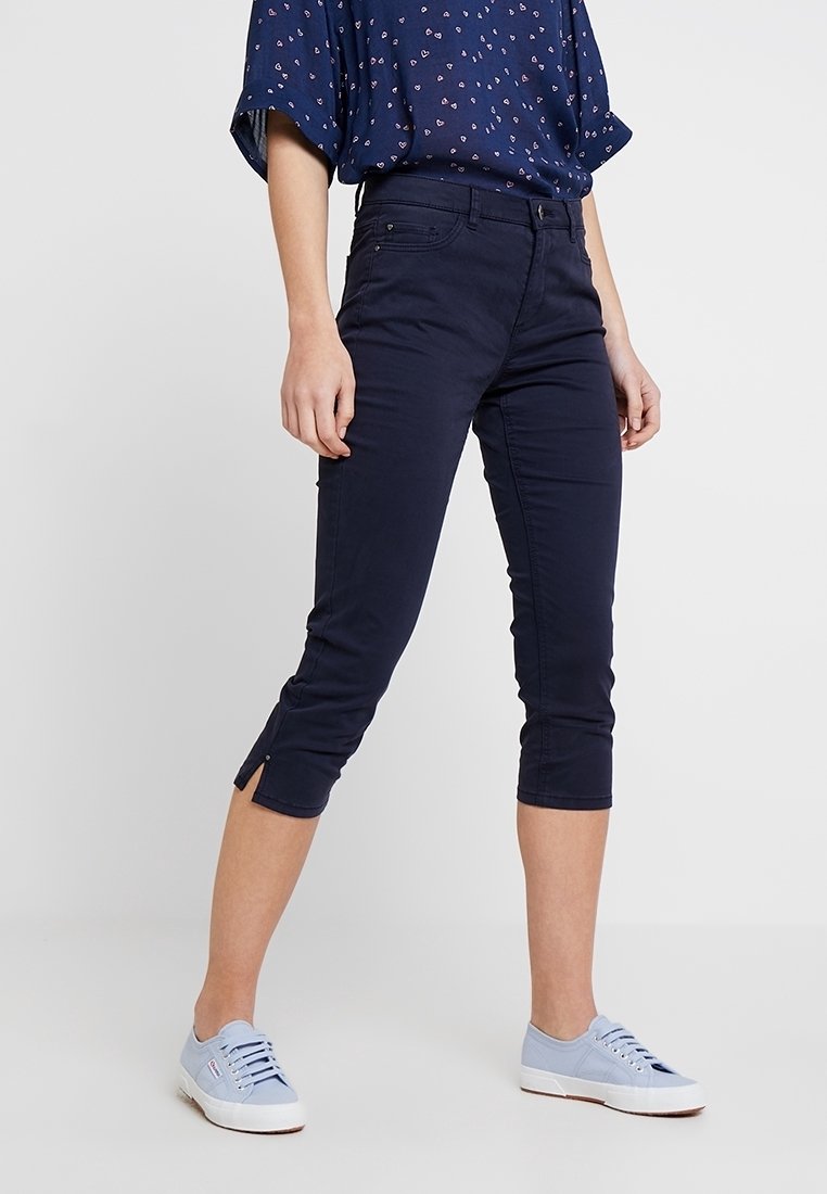 Personne portant un pantalon court bleu marine avec des fentes latérales, un haut bleu à manches courtes à motifs, et des baskets bleu clair, debout sur un fond uni.