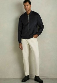 Donkerblauwe bomberjack met een ritssluiting aan de voorkant, geribde manchetten en onderzijde, gecombineerd met crèmekleurige slim-fit broek en zwarte enkellaarzen. Glad van textuur.
