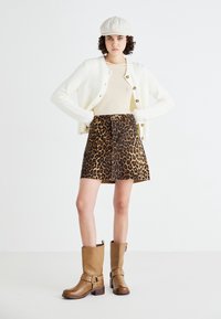 Ung kvinna står med händerna på höfterna, klädd i vit cardigan, beige topp, leopardmönstrad kjol, bruna stövlar och beige keps mot vit bakgrund.